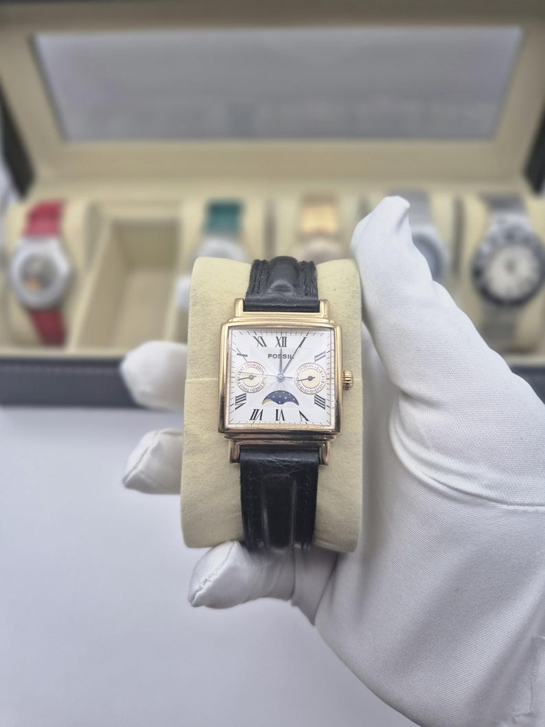 May include: A gold-toned rectangular Fossil watch with a white face, black leather band, and Roman numeral hour markers. The watch features a moon phase indicator and is displayed on a beige cushion.