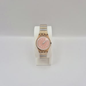 Puede incluir: Un reloj Swatch con esfera rosa y correa de metal dorado. El reloj presenta marcadores de hora con números romanos y un bisel dorado. La correa alterna eslabones dorados y plateados. El reloj se exhibe sobre un soporte blanco.