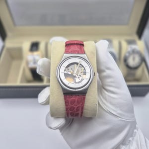 May include: A silver-toned wristwatch with a transparent face revealing the inner workings, set against a cream-colored cushion. It features a red leather band. Other watches are visible in the background within a display case.
