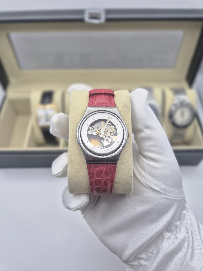 May include: A silver-toned wristwatch with a transparent face revealing the inner workings, set against a cream-colored cushion. It features a red leather band. Other watches are visible in the background within a display case.