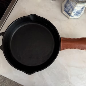 Pode incluir: Uma frigideira de ferro fundido preta com uma capa de couro castanho para a pega. A frigideira é redonda com uma superfície de cozedura plana e uma pequena pega no lado oposto. A capa de couro da pega é cosida e adiciona um toque de estilo.
