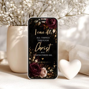 Puede incluir: Funda de teléfono con fondo negro y detalles florales en borgoña y rosa. El texto dorado "I can do all things through Christ who strengthens me" está presente. Un corazón blanco está al lado del teléfono.
