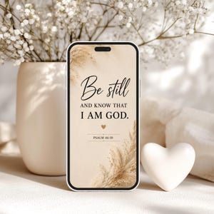 Può includere: Uno smartphone mostra il testo "Be still and know that I am God" su uno sfondo beige con accenti floreali. Un cuore bianco e un vaso di fiori bianchi sullo sfondo.