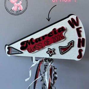 Puede incluir: Decoración blanca en forma de megáfono con ribete negro, con el nombre "Marcia" y "CLASS OF 2026" en rojo y negro. Incluye una estrella, una flecha y las letras "WFHS". La decoración tiene cintas rojas, negras y blancas.