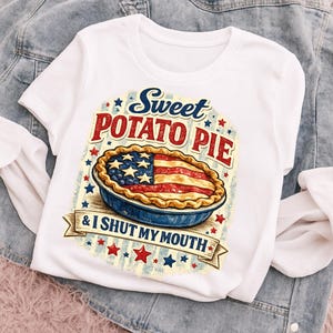 Op de afbeelding: Een wit T-shirt met korte mouwen en een afbeelding van een taart met een Amerikaans vlagpatroon op de korst. De tekst luidt Sweet Potato Pie & I SHUT MY MOUTH, omringd door blauwe en rode sterren op een lichtblauw en wit gestreepte achtergrond.