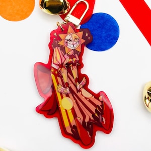 Puede incluir: Un charm de acrílico rojo con un personaje de dibujos animados con cabeza de sol y cara sonriente. El charm tiene un cierre dorado y cuelga de una cinta roja. Campanas doradas y círculos de colores en el fondo.