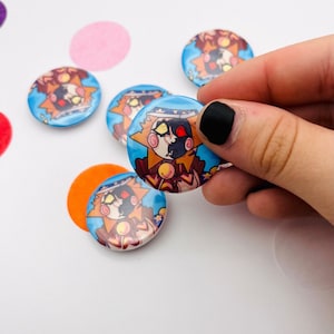 May include: A selection of circular buttons with a cartoon sun character design. The buttons have a blue background and are held by a hand with black nail varnish. Confetti is scattered around the buttons.