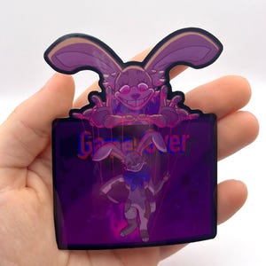 May include: A purple and black sticker featuring a menacing rabbit character with the words "Game Over". The rabbit has a sinister grin and is holding strings, with a smaller rabbit puppet below. The sticker has a glossy finish.