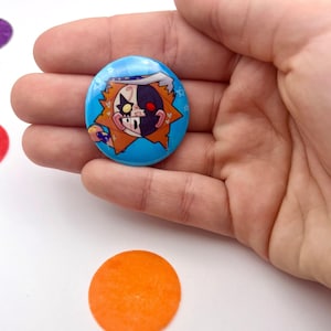 Könnte beinhalten: Ein runder, blauer Button mit einem Cartoon-Charakter mit geteiltem Gesicht. Eine Seite ist weiß und schwarz, die andere schwarz und rot. Der Charakter hat orangefarbene Haare und trägt einen weißen Hut. Der Button wird in einer Hand gehalten.