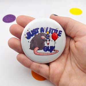 Puede incluir: Un botón blanco con el texto "JUST A LITTLE GUY" en azul, con un dibujo de un opossum con un gorro de fiesta y un globo rojo. El opossum es gris con una cola rosa. Hay confeti esparcido alrededor del botón.