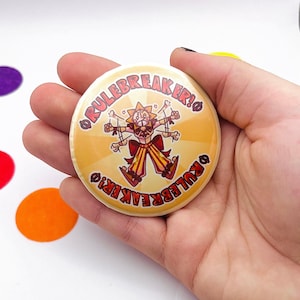 May include: A round, yellow button with the text "RULEBREAKER!" encircling a cartoon character. The character has a sun-like face, multiple arms, and is dressed in red and yellow striped clothing. The button is held in a hand.