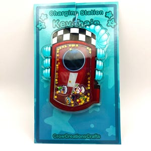 Puede incluir: Un llavero rojo y dorado con el texto "Charging Station Keychain" y una parte superior a cuadros. El llavero tiene un centro transparente con pequeñas figuras de personajes y estrellas doradas. El llavero está en una tarjeta azul con el texto "CrowCreationsCrafts".