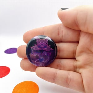 May include: A round, purple button with a spooky ghost design and the text "Spooky Ever". The button has a black background and is held in a hand. The button is a fun accessory.