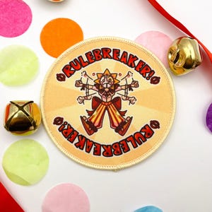 Könnte beinhalten: Ein runder, beigefarbener Aufnäher mit dem Text "RULEBREAKER!" um eine Cartoonfigur herum. Die Figur hat ein sonnenähnliches Gesicht und trägt orangefarbene und gelbe Kleidung. Der Aufnäher ist von bunten Konfetti und goldenen Glocken umgeben.