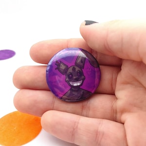 May include: A round button with a purple background featuring a cartoon character with a sinister grin. The character has dark brown fur and large white teeth. The button is held in a person's hand.