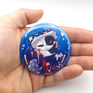 May include: A circular blue badge with a cartoon character design. The character has a white and black face, red eyes, and a white hat with yellow stars. The character is wearing a dark blue outfit with red details. The badge has a glossy finish and white stars.