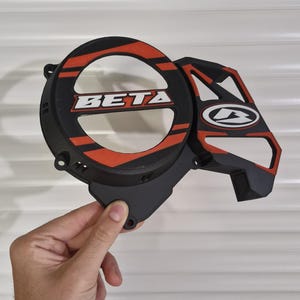 Op de afbeelding: Zwarte en rode motorfiets motordeksel met het woord "BETA" in het wit. De hoes heeft een cirkelvormig ontwerp met rode strepen en een geometrische vorm aan de zijkant. De hoes wordt door een hand vastgehouden.