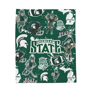 May include: Green and white patterned blanket featuring Michigan State Spartans imagery. The design includes the team's logo, helmet, and mascot, along with the words "Michigan State" and "State". The blanket has a white splatter effect.