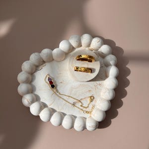 May include: A white marble jewelry tray with a beaded edge holds two gold rings and a gold necklace with colorful gemstones. The tray sits on a light pink surface, showcasing the jewelry.
