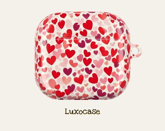 Puede incluir: Una funda blanca para AirPods con un estampado de corazones rojos, rosas y morados. La funda tiene forma redondeada y un pequeño lazo en el lateral. El texto "Luxocase" está impreso en la parte inferior.