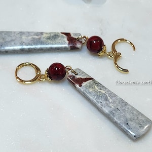 May include: A pair of dangle earrings featuring rectangular gray and burgundy stone pendants. Each earring has a small, round, dark red bead and gold-colored leverback ear wires. The earrings are displayed on a white surface.