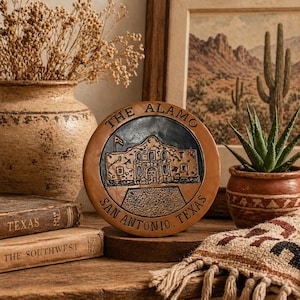 May include: A round, brown ceramic plaque featuring a detailed depiction of The Alamo in San Antonio, Texas. The plaque has the text "THE ALAMO" and "SAN ANTONIO, TEXAS" around the edge. The plaque is displayed on a wooden surface.