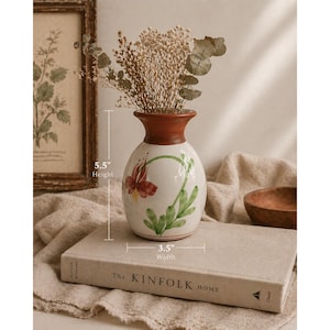 Op de afbeelding: Een witte keramische vaas met een bruine rand en bloemenontwerp, 14 cm hoog en 9 cm breed, bevat gedroogde bloemen en groen. De vaas staat op een boek met de titel "The Kinfolk Home."