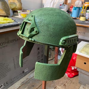 Op de afbeelding: Een groene helm met een getextureerd oppervlak en bevestigde viziercomponenten. De helm is gemonteerd op een houten standaard, die het ontwerp en de constructie laat zien. De algehele esthetiek suggereert een militair of tactisch thema.