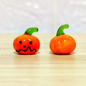 以下が含まれることがあります： 2つのミニチュアガラスカボチャ。カボチャはオレンジ色で、緑色の茎が付いています。1つのカボチャには、黒いジグザグの口と2つの黒い点があります。カボチャは、明るい茶色の木製の表面にあります。