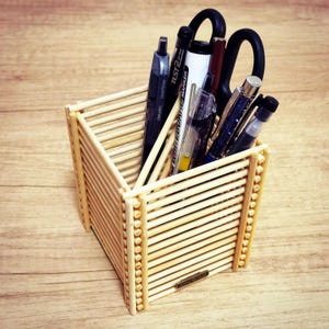 以下が含まれることがあります： 小さな木の棒で作られた手作りの木製デスクオーガナイザー。 ペン、マーカー、ハサミが入っています。 オーガナイザーは、明るい茶色の木製の表面にあります。
