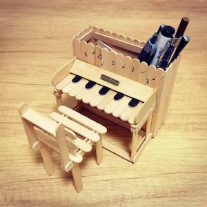 以下が含まれることがあります： 手作りの木製ピアノ型デスクオーガナイザーと、お揃いのミニチュアベンチ。 ピアノは黒い鍵盤と小さな金属製のネームプレートが特徴です。 オーガナイザーにはペンや鉛筆が入っています。 側面には音符がエッチングされています。