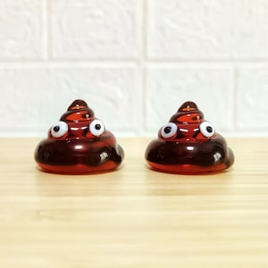 May include: Two small, dark brown, swirl-shaped glass figurines, each featuring a pair of white and black googly eyes attached to the front. The glass has a smooth, glossy finish and a translucent quality, reflecting light on the rounded surfaces.