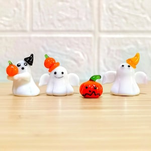 以下が含まれることがあります： ハロウィーンをテーマにしたガラス製の置物4つ。黒、オレンジ、黄色の帽子をかぶった3つの白いゴーストのフィギュアと、笑顔のオレンジ色のカボチャ。ゴーストのフィギュアはカボチャを持ったり、身につけたりしています。フィギュアは明るい茶色の表面にあります。