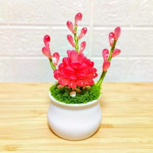 以下が含まれることがあります： 小さな白いセラミックポットに、鮮やかな造花のアレンジメントが入っています。アレンジメントは、明るい赤色の花びらの中心的なクラスターと、緑色の茎に繊細なピンク色のつぼみがあります。ポットは木製の表面に置かれています。