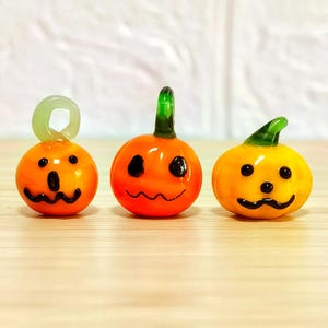 Pode incluir: Três pequenas abóboras de vidro laranja com traços faciais pretos e caules verdes. As abóboras têm um acabamento brilhante e estão dispostas sobre uma superfície de madeira clara. Decoração festiva para o Halloween.