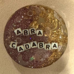May include: A round, resin pendant with the words "ABRA CADABRA" spelled out in white letter tiles. The pendant has a glittery, maroon base with gold leaf accents and a clear, glossy finish.