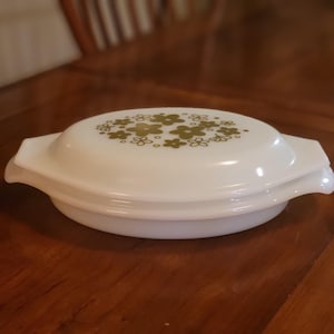 May include: White oval casserole dish with a lid featuring a pattern of green flowers. The dish has handles on either side and is sitting on a wooden surface. A vintage kitchenware item.