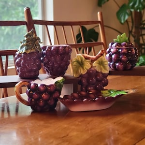 May include: A collection of ceramic grape-themed kitchenware. The set includes a creamer, a covered jar, a small dish, and two decorative containers, all in shades of purple and green. The items are arranged on a wooden surface.