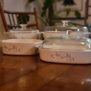 May include: A set of four off-white casserole dishes with glass lids. Each dish features a delicate floral design in shades of purple and green. The dishes are arranged on a wooden table, showcasing their classic design.