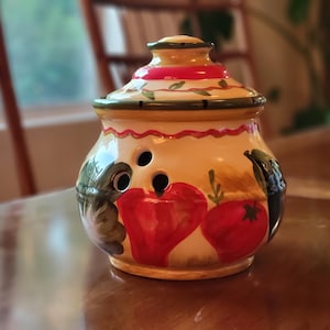 May include: A ceramic garlic keeper with a lid. The jar is painted with red tomatoes, green peppers, and other vegetables. The lid has a red and green band. The jar has three holes for ventilation.