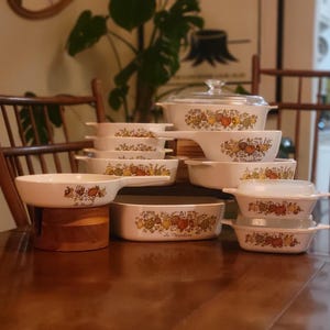 May include: A collection of vintage white ceramic cookware with a colourful floral and fruit design. The set includes various sizes of casserole dishes, baking dishes, and a skillet. The cookware is arranged on a wooden table, showcasing its classic design.