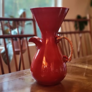 May include: A vibrant red ceramic jug with a flared top and a rounded base. It has a small spout and a woven handle. This jug is designed for serving drinks and adds a splash of colour to any table. It is likely used for serving beverages.