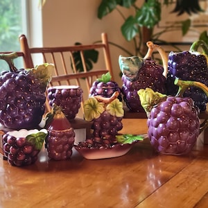 Puede incluir: Una colección de piezas de servicio de cerámica con temática de uvas. El conjunto incluye una tetera, una crema, un azucarero y fuentes para servir, todo en tonos morados y verdes. Las piezas están dispuestas sobre una mesa de madera.