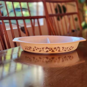 May include: Cream-colored oval divided serving dish with brown floral pattern. The dish has two compartments and handles on each end. The dish is sitting on a wooden table.