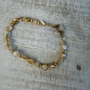 May include: A beaded bracelet featuring iridescent, heart-shaped beads in shades of cream and gold. The bracelet is strung on a gold-colored chain and has a gold-colored clasp.