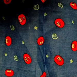 May include: Denim fabric with a repeating pattern of red cartoon faces, green stars, and green spiral designs. The fabric has a blue background and is likely used for clothing or home decor.