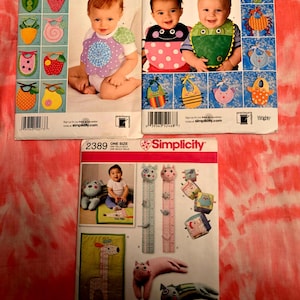 May include: Three Simplicity sewing pattern envelopes featuring baby bibs and toys. The bib patterns include various designs like fruits, animals, and floral patterns. The toy patterns include a giraffe, cat, and bear. The text "Simplicity" and "ONE SIZE" are visible.