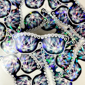 Op de afbeelding: Een verzameling holografische stickers met een zonnebril met de tekst "LASER THERAPY" en het hashtag-symbool. De zonnebril heeft een zwart montuur en een ontwerp dat licht reflecteert in een regenboogpatroon.