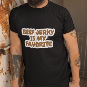 Op de afbeelding: Zwart T-shirt met de tekst "BEEF JERKY IS MY FAVORITE" in een witte en bruine letter. Het shirt wordt gedragen door een persoon met een baard en tatoeages. De persoon draagt een blauwe pet en een zwarte broek.