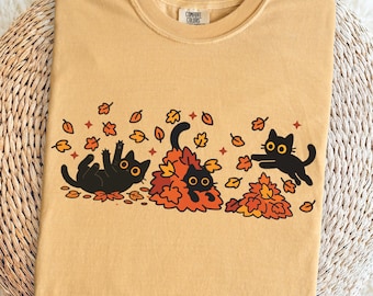 Puede incluir: Camiseta de color mostaza con un gráfico de tres gatos negros jugando entre hojas de otoño. Un gato rueda, otro está en un montón de hojas y el tercero alcanza una hoja. La camiseta tiene cuello redondo y la marca "Comfort Colors".